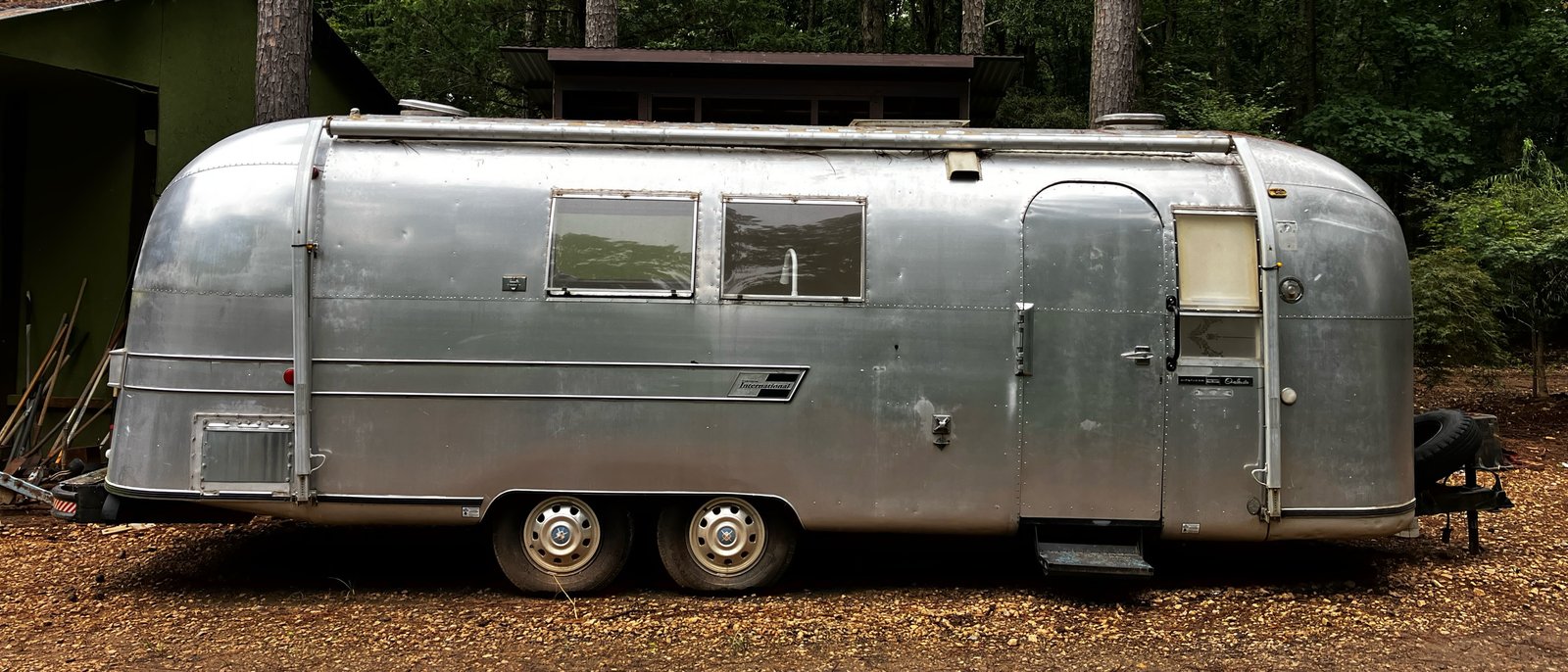 The PrayerStream: a 1967 Airstream travel trailer, seen side-on, parked on gravel among tall pines