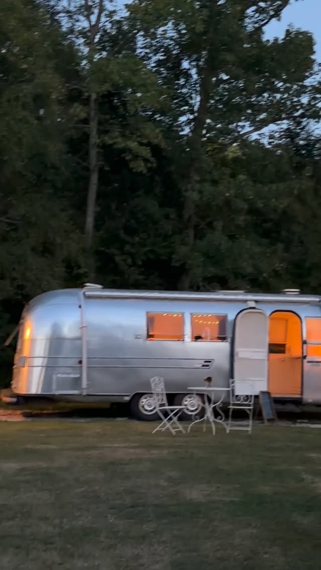 Reel: the Airstream at dusk, lit inside, with chairs set out in front
