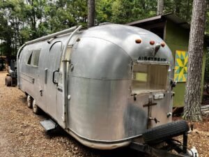 The Airstream from the back corner, parked on gravel beside a green shed with a painted quilt block